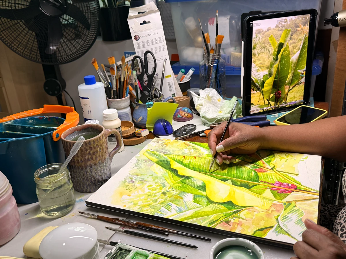 Sharon’s hands painting tropical leaves, with brushes and paints on the studio table.